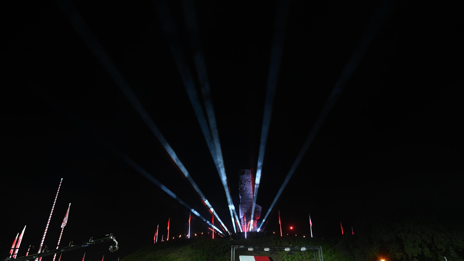 Das mit Lichtern und einer polnischen Flagge geschmückte Kriegerdenkmal Westerplatte, davor eine militärische Ehrengarde und ein Rednerpult. Das mit Lichtern und einer polnischen Flagge geschmückte Kriegerdenkmal Westerplatte, davor eine militärische Ehrengarde und ein Rednerpult.