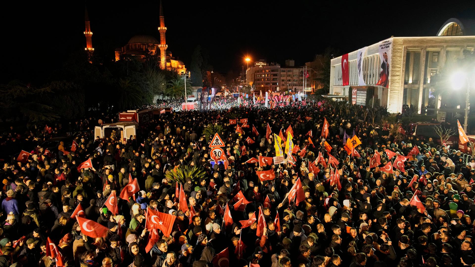 Viele Menschen versammeln sich vor einem Gebäude und schwenken teilweise rote Fahnen.