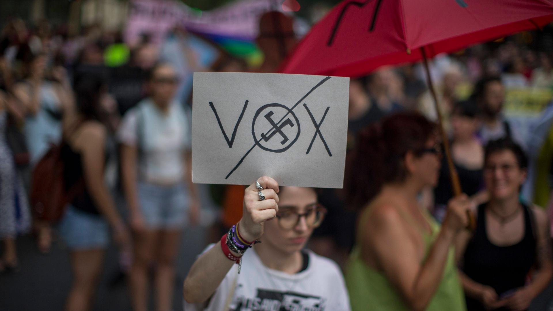 Bei einer Demonstration in Madrid hält eine junge Frau mit eiener Hand ein Plakat mit der Aufschrift VOX in die Höhe. Im Buchstaben O ist ein Hakenkreuz eingezeichnet und dieses ist durchgestrichen.