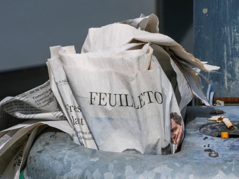Eine gedruckte Zeitung steckt im Mülleimer einer Haltestelle. Aufgeschlagen ist das Feuilleton.