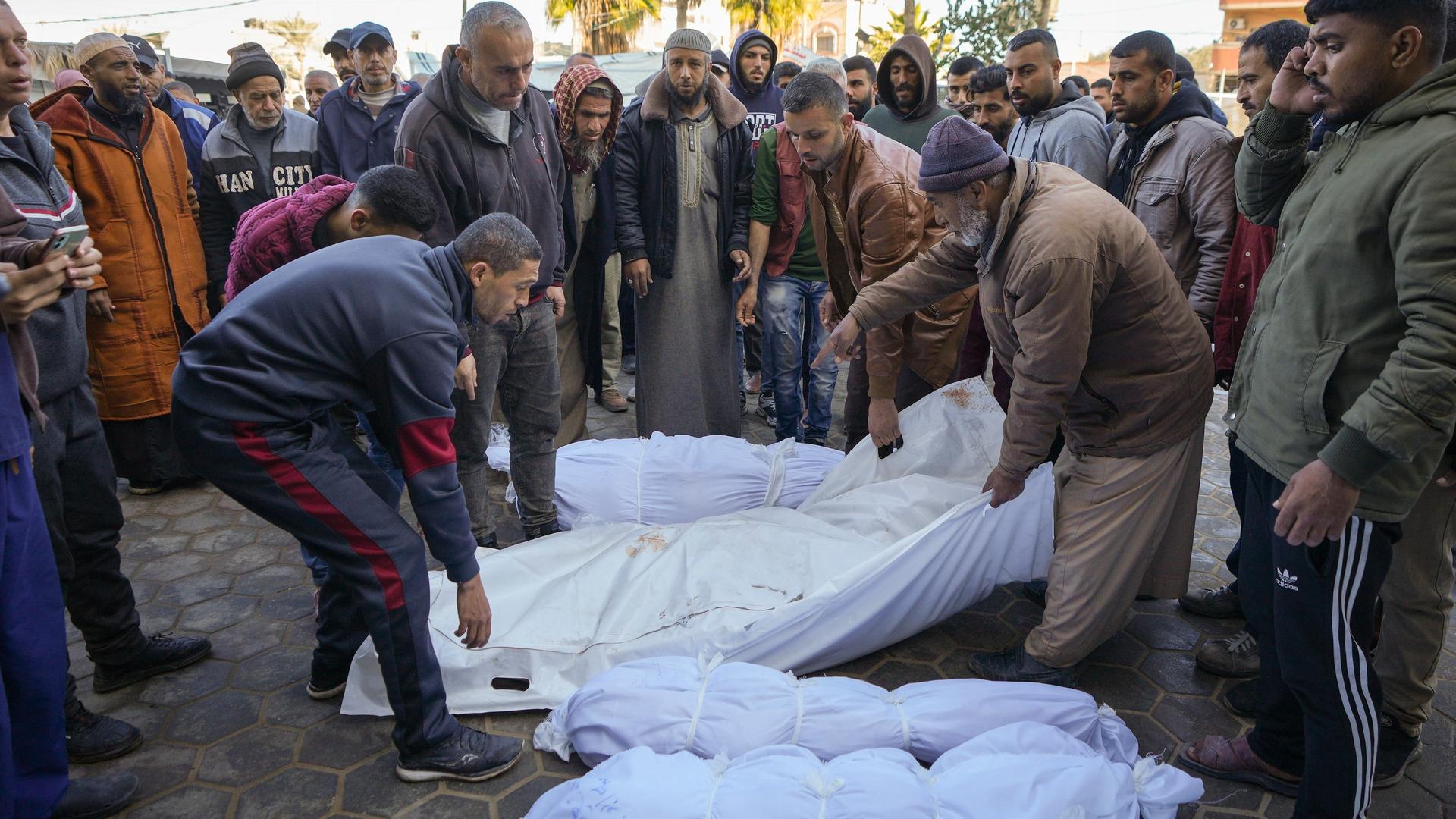 Menschen im Gazastreifen stehen im Halbkreis um Leichen, die in Tücher gewickelt sind. 