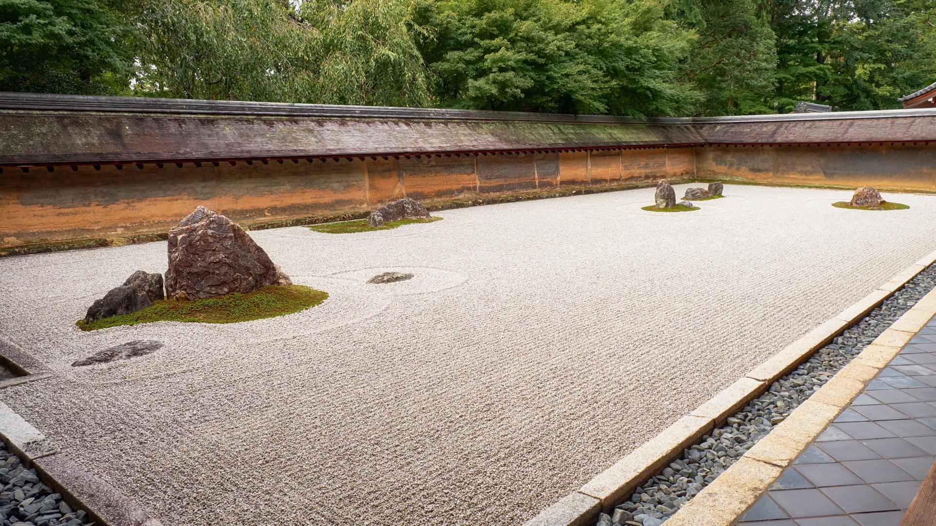 Ein Rechteck auf dem Boden, das mit Sand gefüllt ist, der ordentlich geharkt wurde. Im Sand sind Steine angeordnet. Es ist ein Steingarten am Ryōan-ji Tempel in Kyoto.