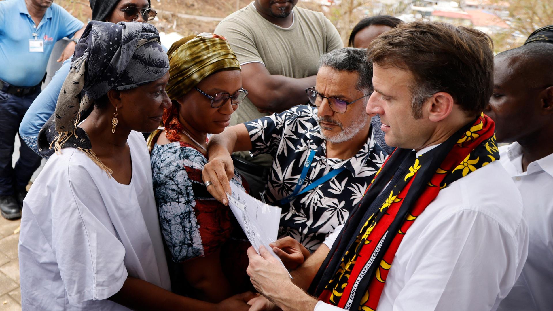 Der französische Präsident Emmanuel Macron hält ein Blatt Papier in der Hand. Vor ihm stehen zwei Frauen aus dem Überseegebiet Mayotte. Der französische Präsident Emmanuel Macron hält ein Blatt Papier in der Hand. Vor ihm stehen zwei Frauen aus dem Überseegebiet Mayotte.