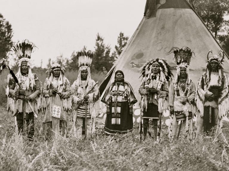 Jun. 06, 1913 - USA - Group of Siksika (Blackfeet) men and one woman singing in front of tipi - Moki rain maker and chief in costume, World's Fair, St. Louis, 1904