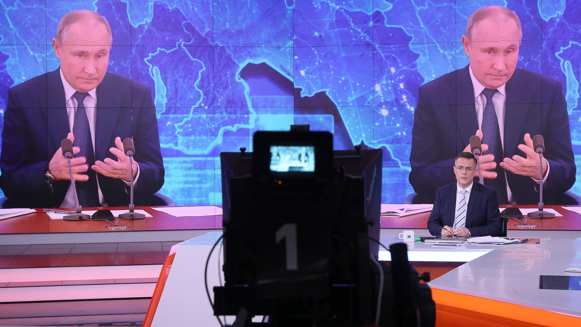 Auf einer großen Monitorwand ist zweimal Putin zu sehen, der seine Hände lose vor sich hält. Klein vor ihm sitzt ein Moderator und steht eine Kamera.