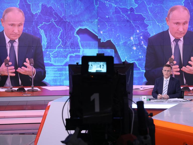 Auf einer großen Monitorwand ist zweimal Putin zu sehen, der seine Hände lose vor sich hält. Klein vor ihm sitzt ein Moderator und steht eine Kamera.