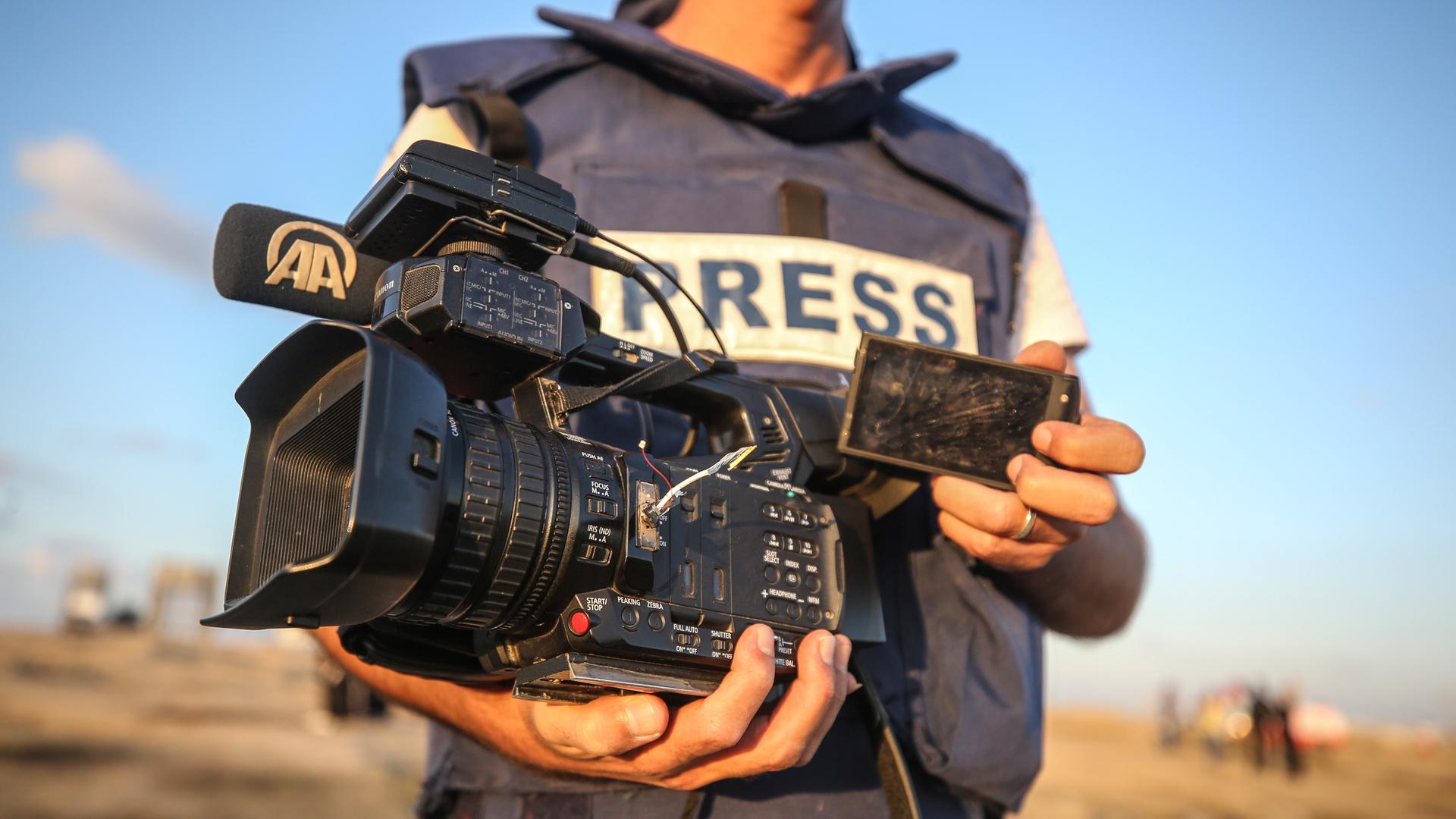 Nahaufnahme eines Mannes in einer kugelsicheren Weste mit der Aufschrift „PRESS“. In der Hand hält er eine defekte Kamera. Nahaufnahme eines Mannes in einer kugelsicheren Weste mit der Aufschrift „PRESS“. In der Hand hält er eine defekte Kamera.