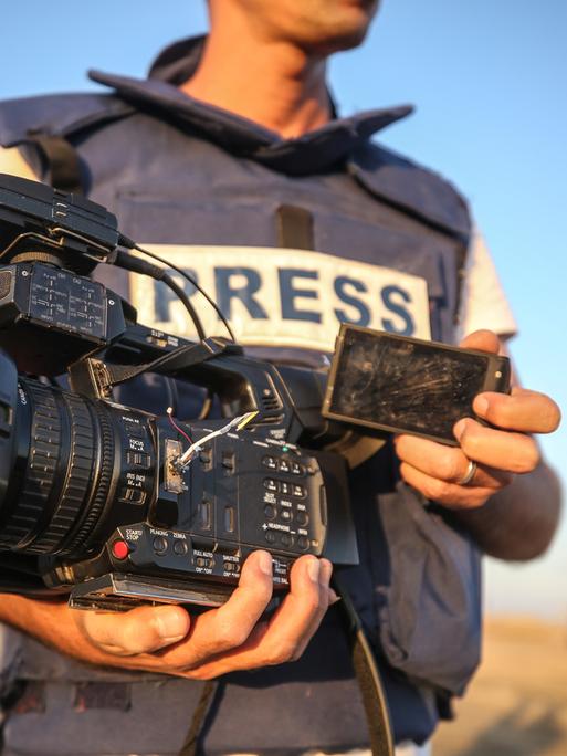 Nahaufnahme eines Mannes in einer kugelsicheren Weste mit der Aufschrift „PRESS“. In der Hand hält er eine defekte Kamera.