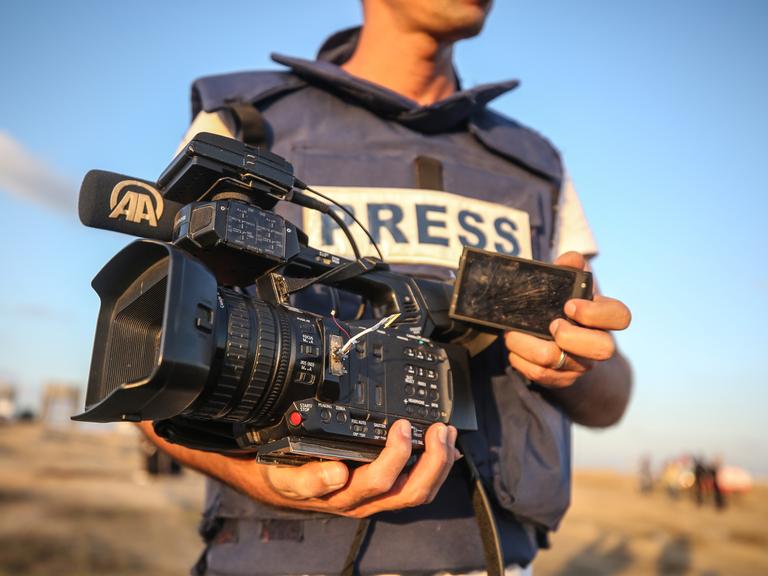 Nahaufnahme eines Mannes in einer kugelsicheren Weste mit der Aufschrift „PRESS“. In der Hand hält er eine defekte Kamera. Nahaufnahme eines Mannes in einer kugelsicheren Weste mit der Aufschrift „PRESS“. In der Hand hält er eine defekte Kamera.