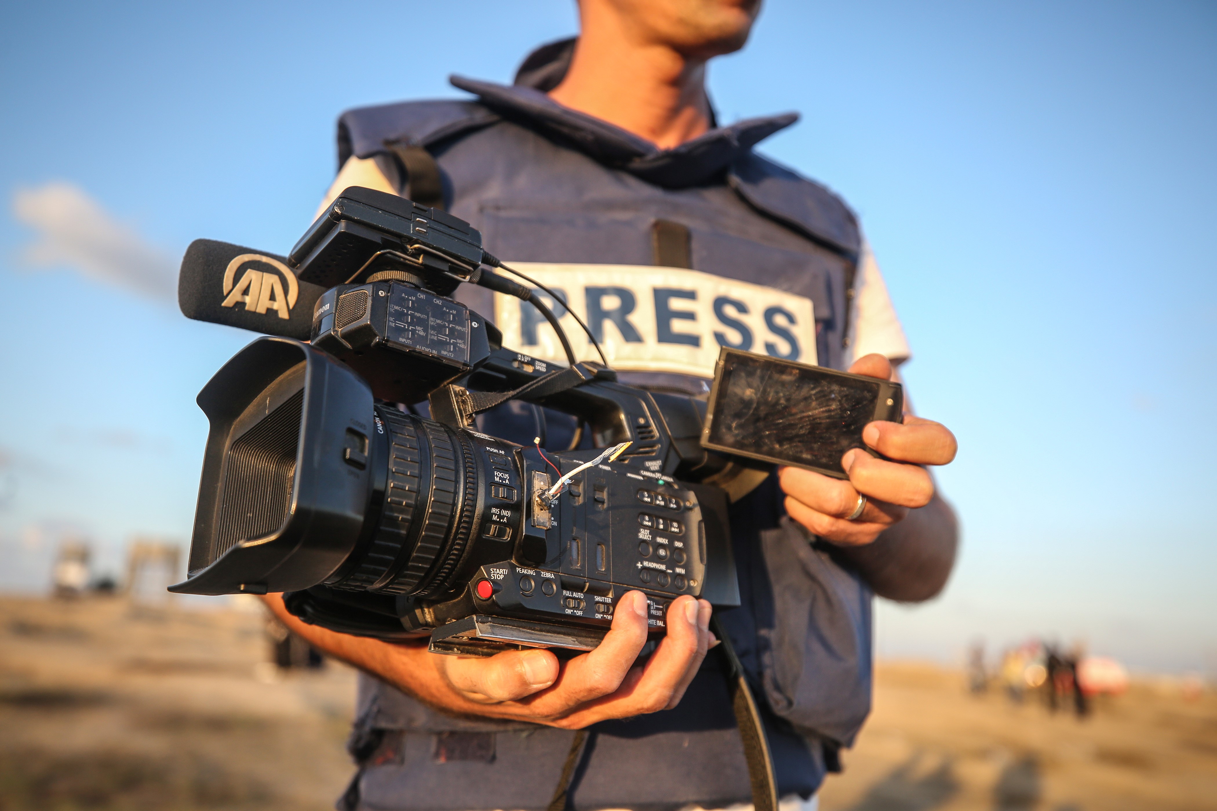 Nahaufnahme eines Mannes in einer kugelsicheren Weste mit der Aufschrift „PRESS“. In der Hand hält er eine defekte Kamera.
