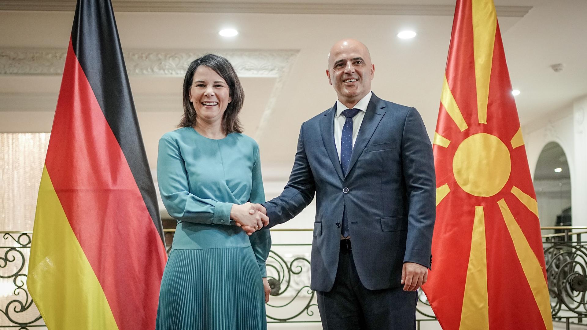 Annalena Baerbock und Dimitar Kovahevski geben sich die Hand. Beide lachen. Neben den Politikern steht jeweils die deutsche und die nordmazedonische Flagge.