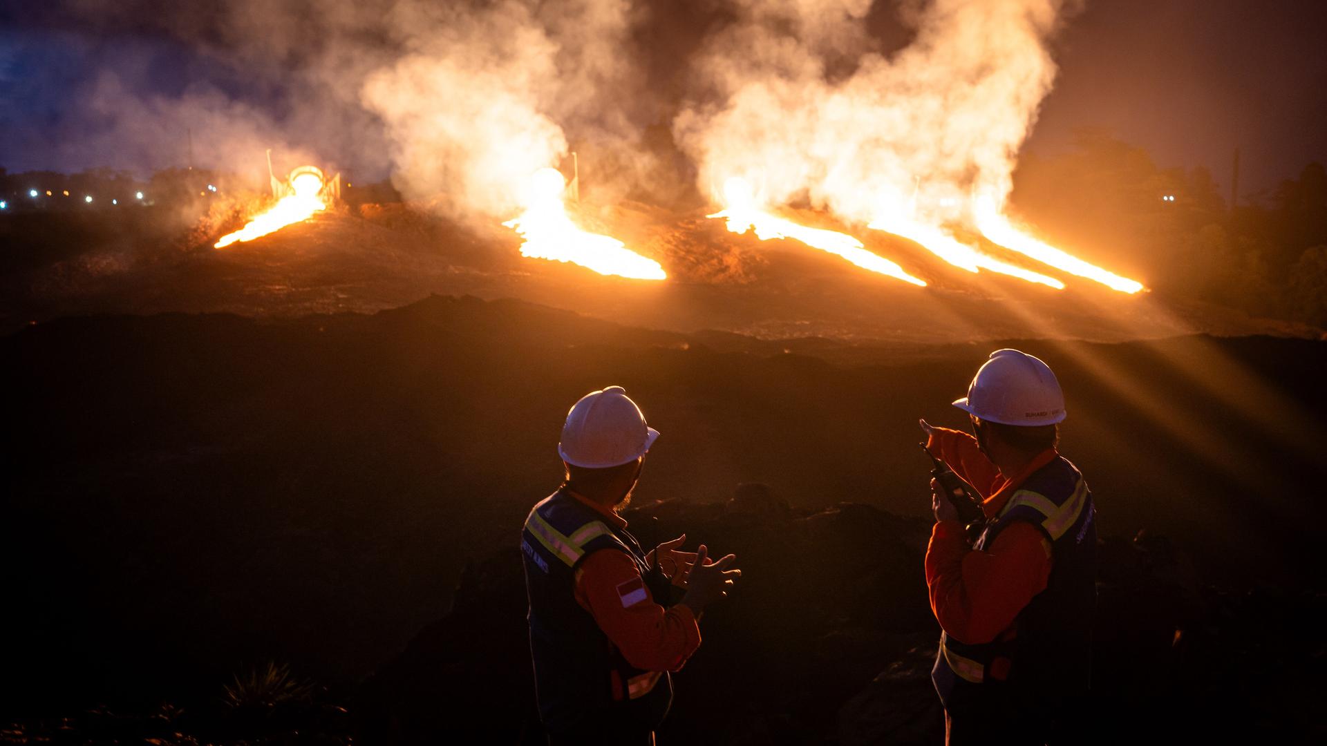 Nickelproduktion in Indonesien. Zwei Wachposten mit weißen Helmen und Feuerschutzwesten beobachten gleißendes Licht auf einer Mine. Das Licht ist in Orange getaucht.