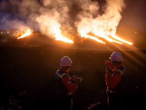 Nickelproduktion in Indonesien. Zwei Wachposten mit weißen Helmen und Feuerschutzwesten beobachten gleißendes Licht auf einer Mine. Das Licht ist in Orange getaucht.