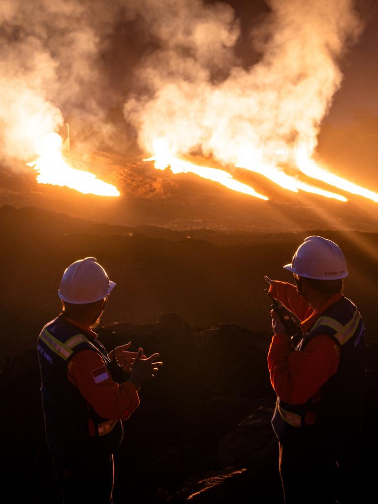 Nickelproduktion in Indonesien. Zwei Wachposten mit weißen Helmen und Feuerschutzwesten beobachten gleißendes Licht auf einer Mine. Das Licht ist in Orange getaucht.