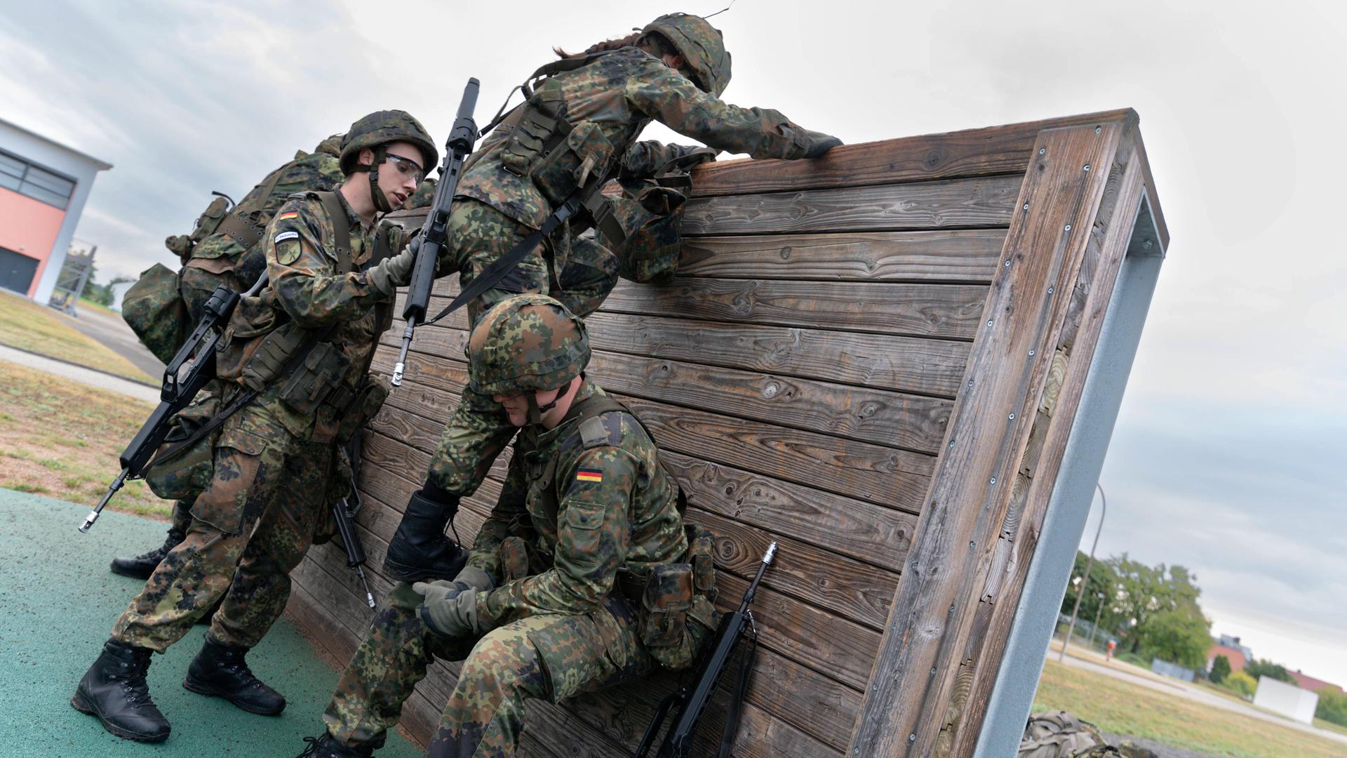 3 Soldaten in Uniformen und mit Helmen und Gepäck helfen sich beim Klettern über eine Holz-Wand.