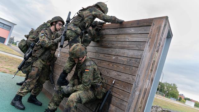 3 Soldaten in Uniformen und mit Helmen und Gepäck helfen sich beim Klettern über eine Holz-Wand.