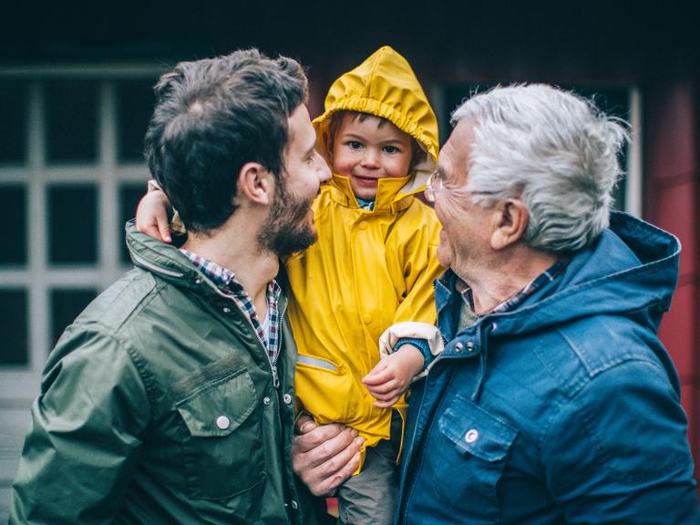 Ein Vater und ein Großvater tragen einen kleinen Jungen auf dem Arm - der Junge lacht in die Kamera, die beiden Erwachsenen lachen den Jungen an.
