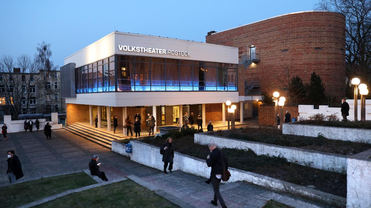 Blick auf das weiße modern wirkende Gebäude mit Pflanzen-Terrassen. Davor laufen und sitzen Menschen.