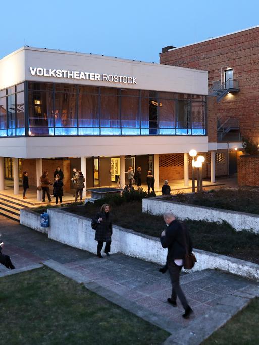 Blick auf das weiße modern wirkende Gebäude mit Pflanzen-Terrassen. Davor laufen und sitzen Menschen.