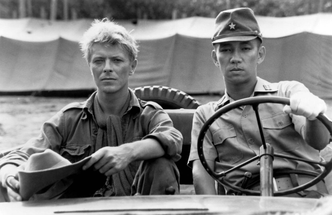 Die Filmszene in Schwarz-Weiß zeigt David Bowie (links) und Ryūichi Sakamoto (rechts) in einem Auto sitzend.