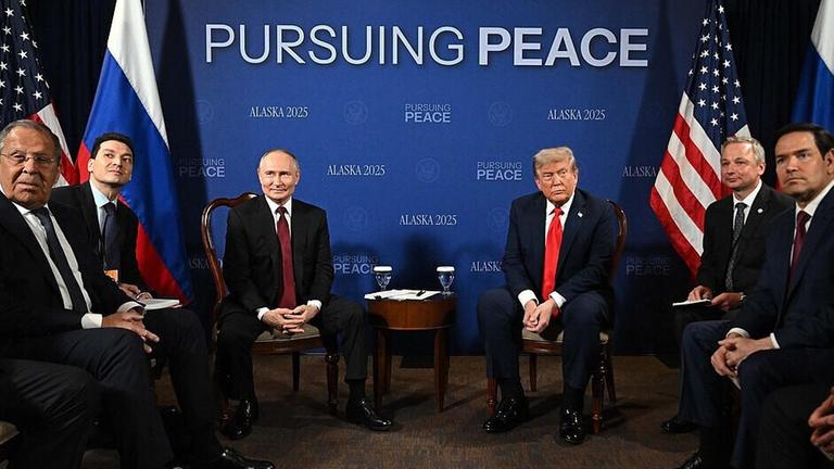 Wladimir Putin und Donald Trump sitzen nebeneinander auf Stühlen. Zwischen ihnen ist ein kleiner Tisch. Hinter ihnen eine blaue Wand mit den Worten "Alaska 2025" und auf englisch "Den Frieden verfolgen". 