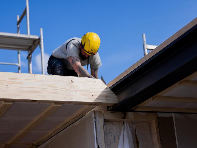 Ein Dachdecker mit gelbem Schutzhelm sitzt auf dem Dach eines Fertighauses.