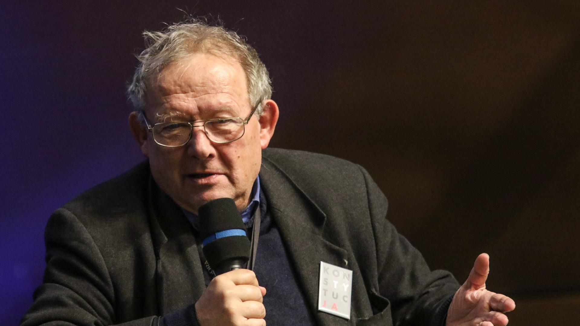 Der ehemalige polnische Dissident Adam Michnik hält sich ein Mikrofon vor das Gesicht und gestikuliert mit der linken Hand.