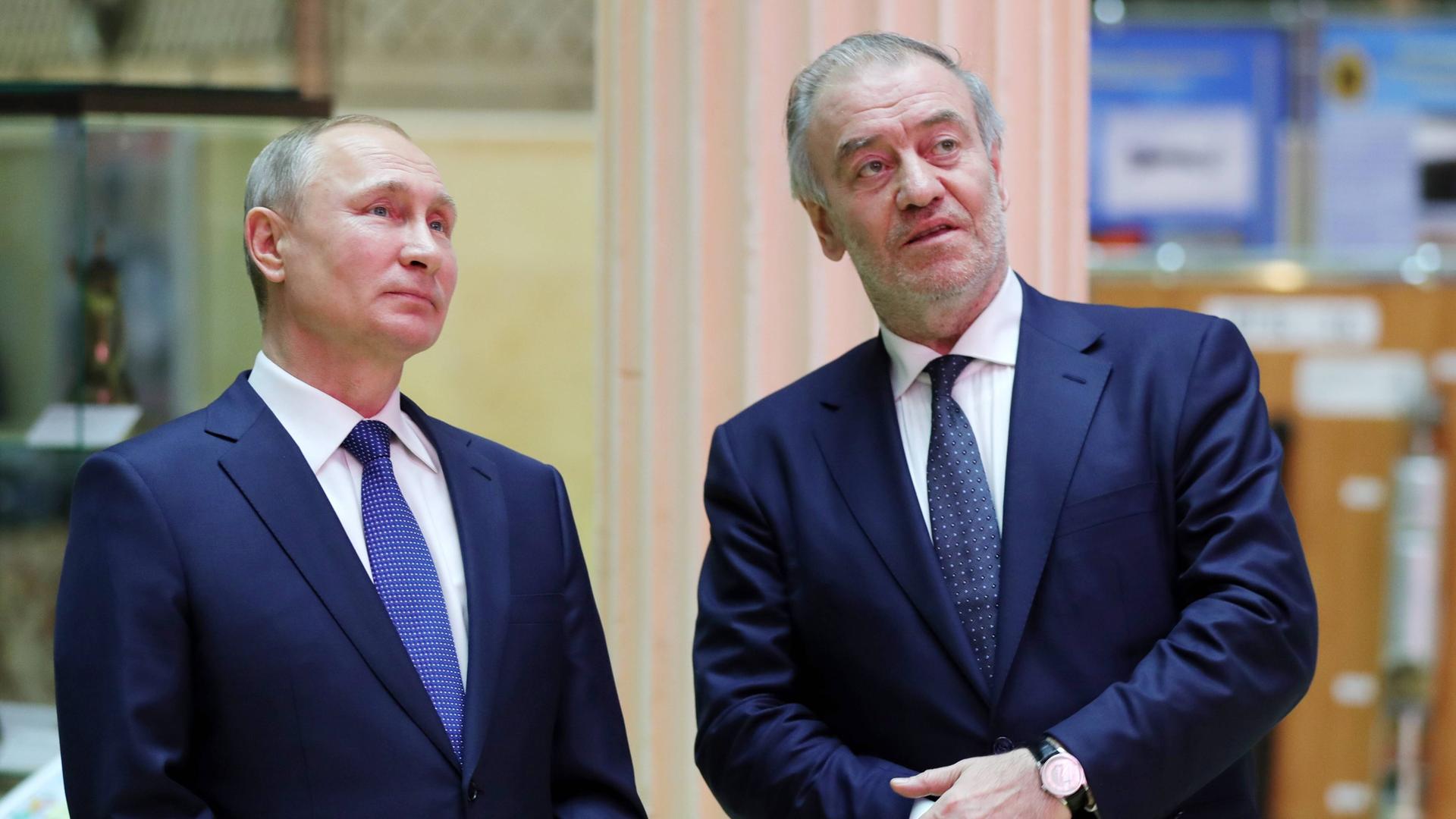 Der russische Präsident Wladimir Putin und der Dirigent Waleri Gergijew stehen nebeneinander. Putin auf der linken Seite, Gergijew auf der rechten. Sie tragen beide dunkelblaue Anzüge.