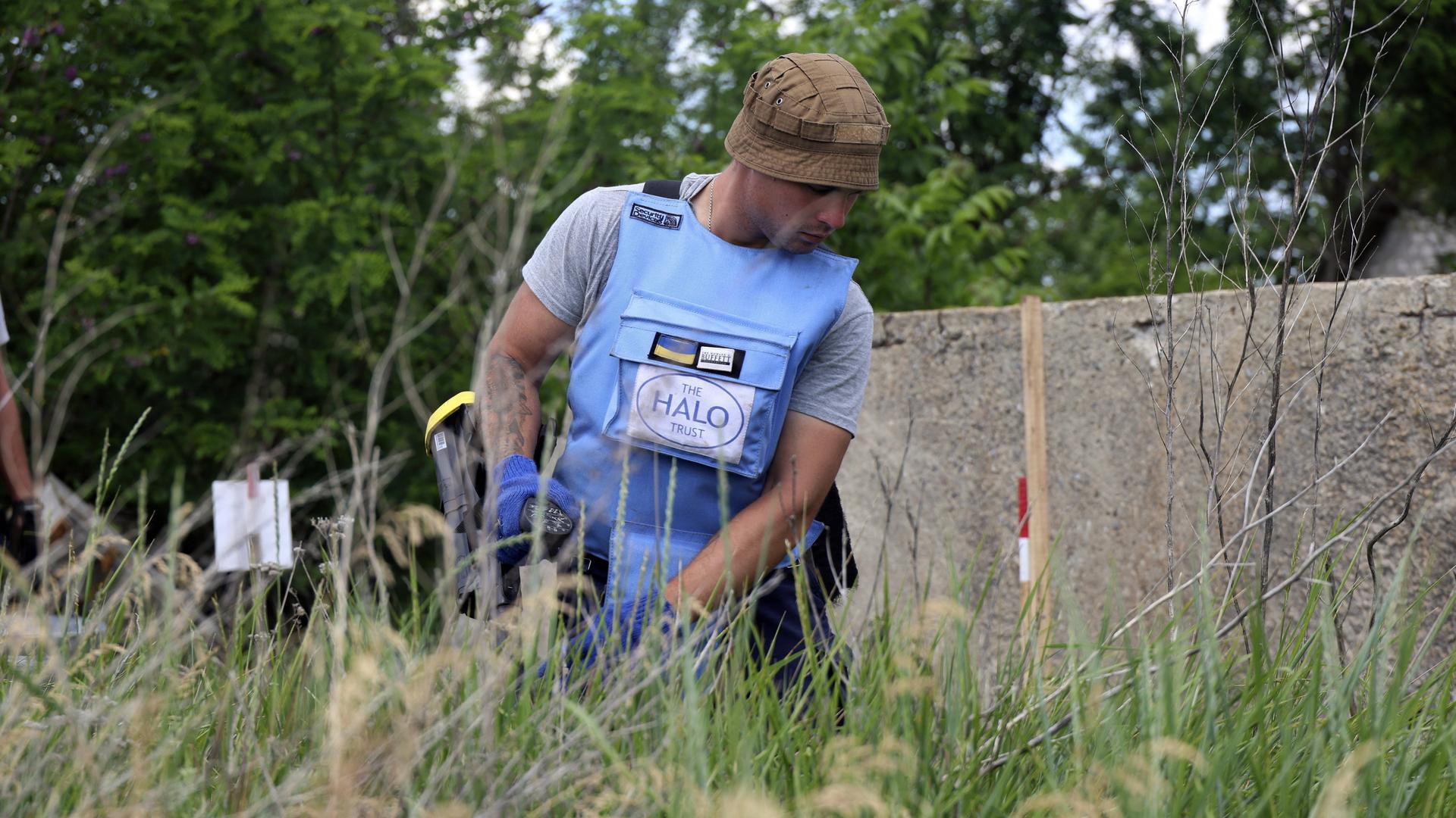 ein Mann in einer hellblauen Splitterschutzweste kniet im Gras. er ist ukrainischer Minenräumer und durcsucht eine Wiese nahe Charkiw.