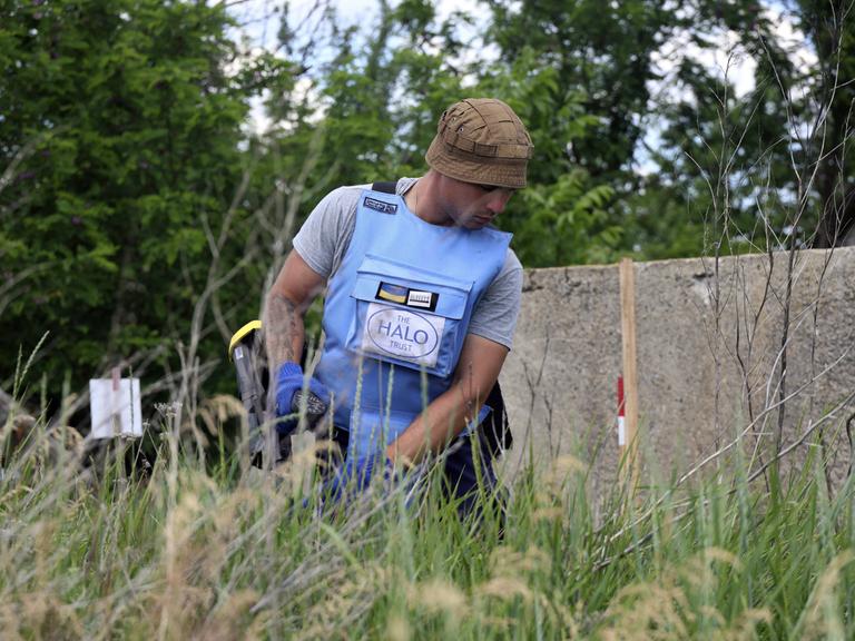 ein Mann in einer hellblauen Splitterschutzweste kniet im Gras. er ist ukrainischer Minenräumer und durcsucht eine Wiese nahe Charkiw.