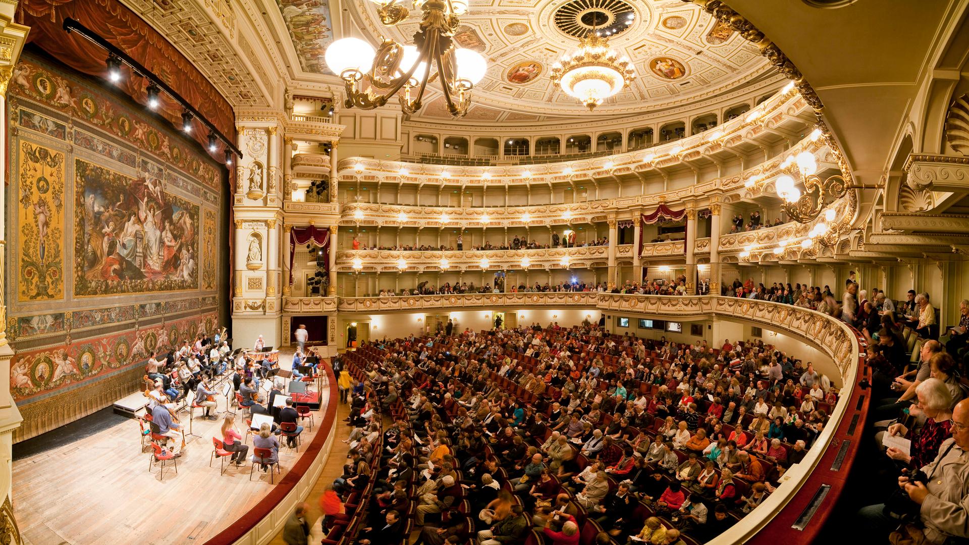 Die Semperoper in Dresden von innen, ein voll besetzter Saal mit Publikum und Musikern auf der Bühne