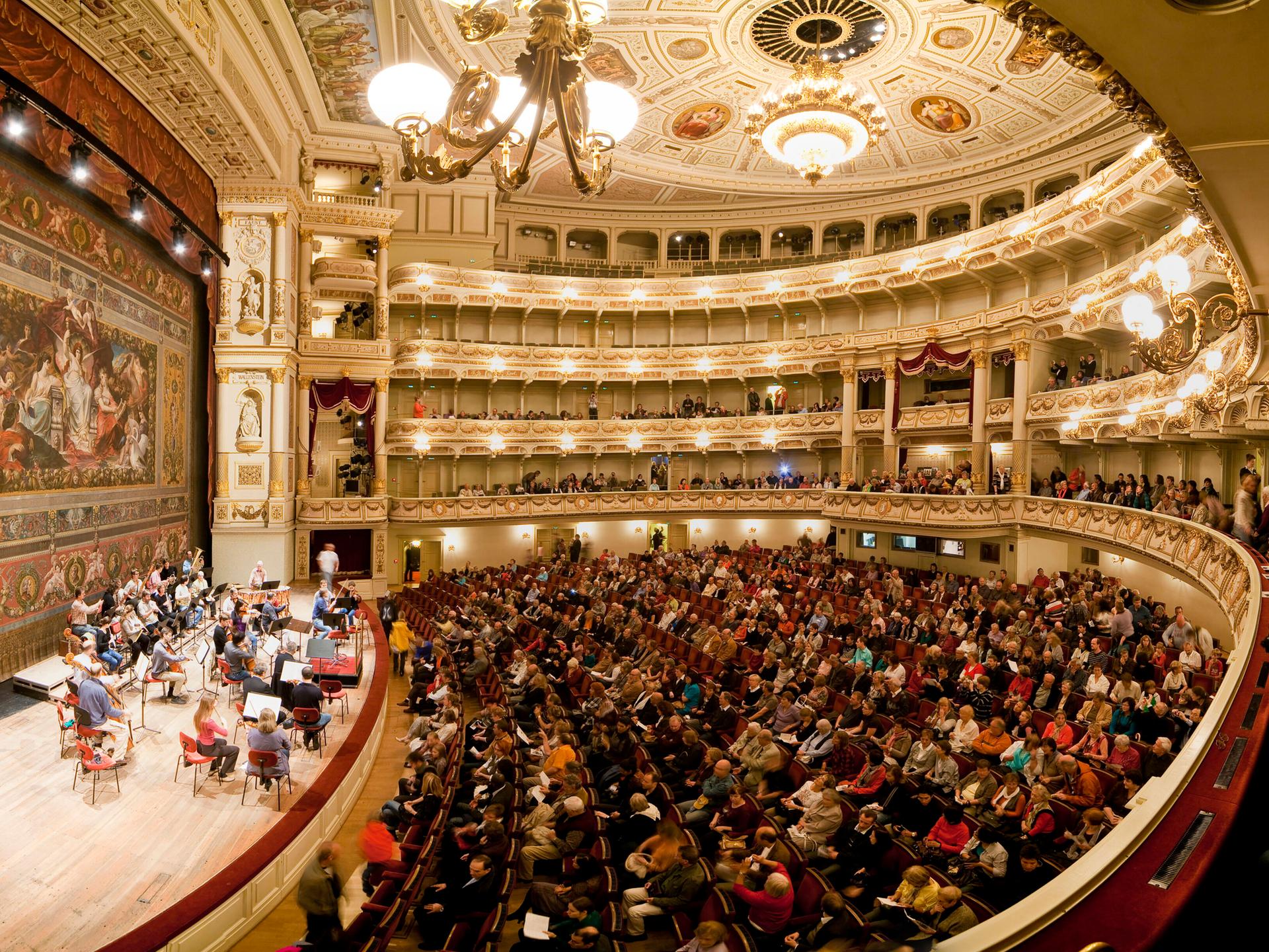 Die Semperoper in Dresden von Innen, ein voll besetzter Saal mit Publikum und Musikern auf der Bühne. 