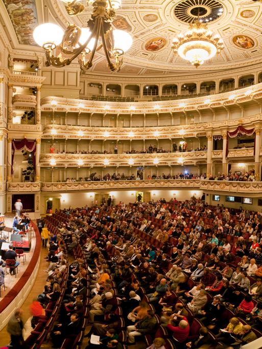 Die Semperoper von Innen, ein voll besetzter Saal mit Publikum und Musikern auf der Bühne. 