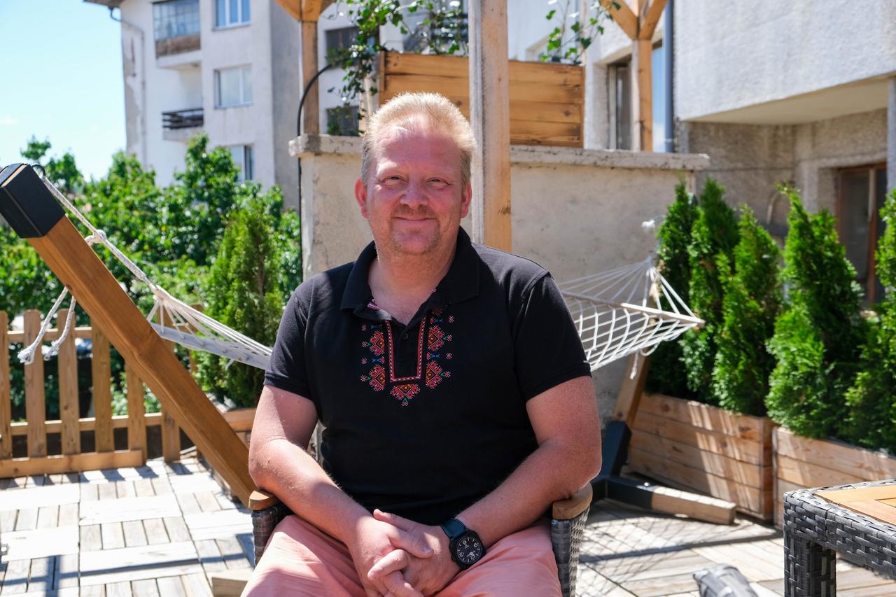 Ein Mann mit Dreitagebart und T-Shirt sitzt auf einer Terrasse.