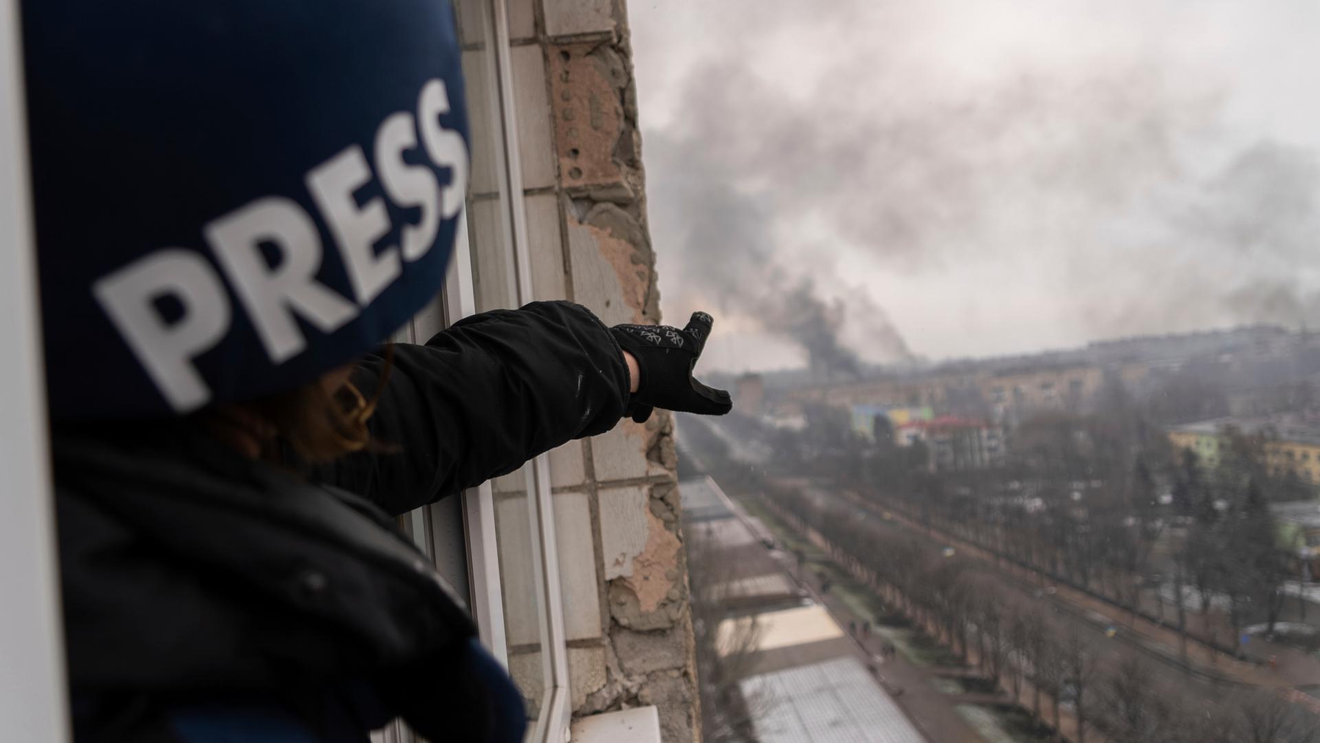 Ein Journalist mit einem Helm und der Aufschrift "Press" steht in einem zerstörten Haus und zeigt auf eine Rauchwolke in der Ferne.