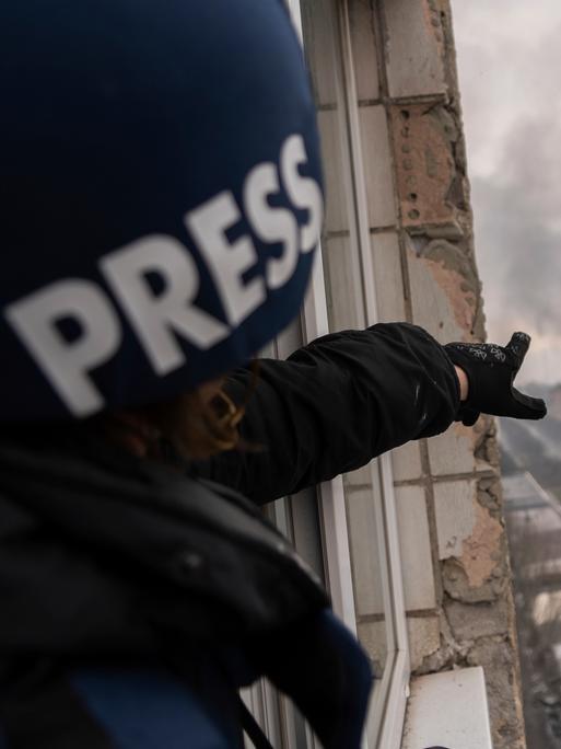 Ein Journalist mit einem Helm und der Aufschrift "Press" steht in einem zerstörten Haus und zeigt auf eine Rauchwolke in der Ferne.