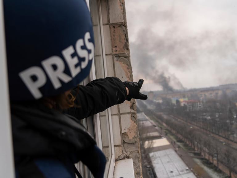 Ein Journalist mit einem Helm und der Aufschrift "Press" steht in einem zerstörten Haus und zeigt auf eine Rauchwolke in der Ferne.