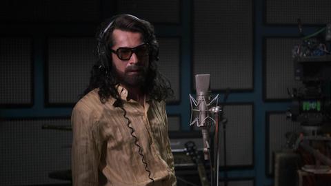 Im Still aus "Cem Karaca" steht der Protagonist mit langen Haaren und in einem seidenen Hemd in einem Aufnahmestudio neben dem Mikrofon.