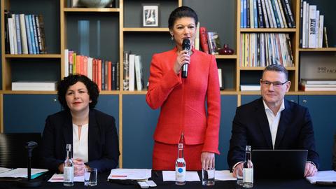 Sahra Wagenknecht (M) begrüßt zusammen mit Amira Mohamed Ali (l) und Ralph Suikat die Teilnehmer zum Gründungsakt der Partei "Bündnis Sahra Wagenknecht - für Vernunft und Gerechtigkeit". Vor ihnen ein Tisch mit mehreren Wasserflaschen, hinter den Politikern eine Wand mit Büchern.