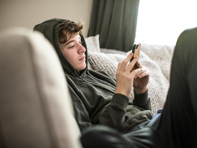 Ein Teenager sitzt auf der Couch und schaut auf sein Smartphone.