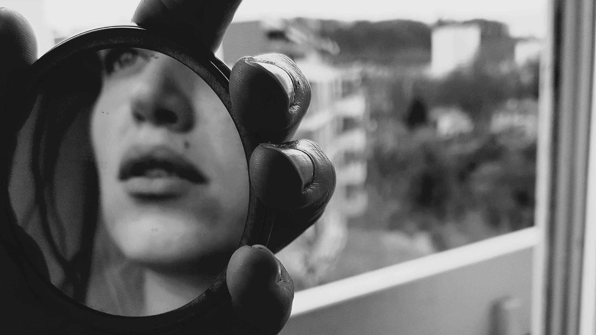 Schwarz-Weiß-Bild einer Hand, die einen Handspiegel hält, darin ist ansatzweise das Gesicht einer jungen Frau zu erkennen. Die junge Frau steht in einer Wohnung vor dem Fenster.