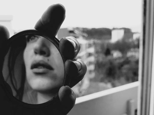 Schwarz-Weiß-Bild einer Hand, die einen Handspiegel hält, darin ist ansatzweise das Gesicht einer jungen Frau zu erkennen. Die junge Frau steht in einer Wohnung vor dem Fenster.