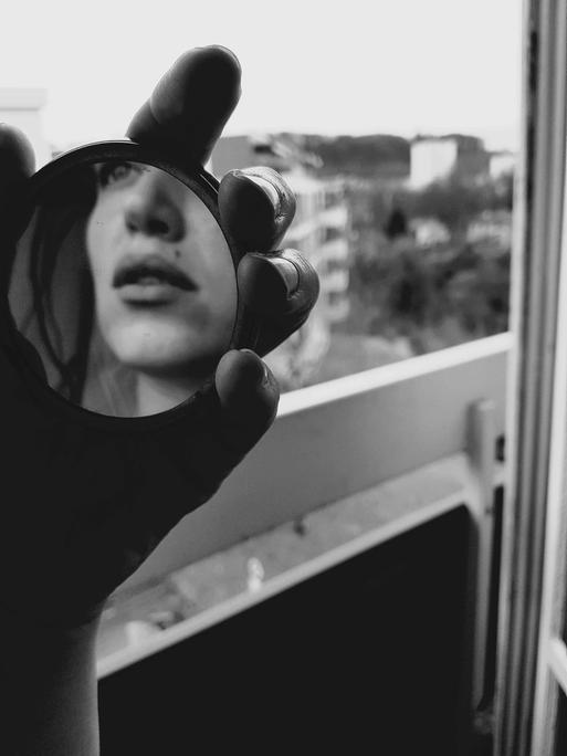 Schwarz-Weiß-Bild einer Hand, die einen Handspiegel hält, darin ist ansatzweise das Gesicht einer jungen Frau zu erkennen. Die junge Frau steht in einer Wohnung vor dem Fenster.