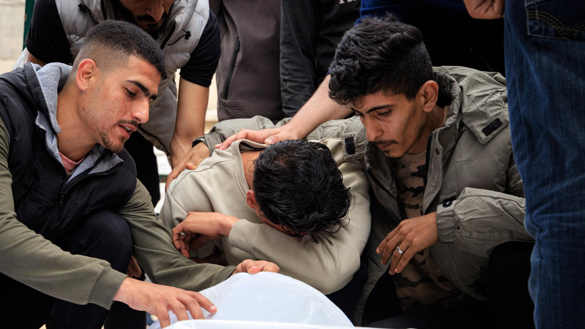 Palästinenser trauern um ihre Angehörigen, die bei erneuten Angriffen in Gaza-Stadt ums Leben gekommen sind. Zu sehen sind drei Männer, die um einen zugedeckten Leichnam sitzen. Einer von ihnen hat das Gesicht in seinen Armen vergraben. 