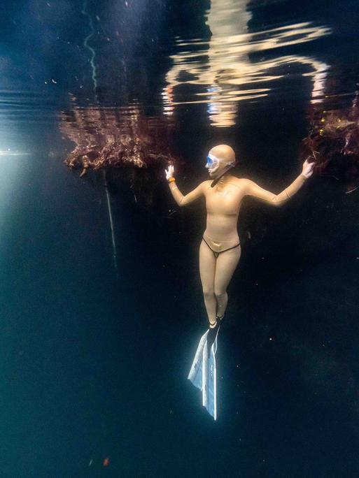 Eine Taucherin steht förmlich unter Wasser. Sie trägt einen hellorangen Tauchanzug und hält sich mit beiden Händen an undeutlichen, schwimmenden Strukturen fest.