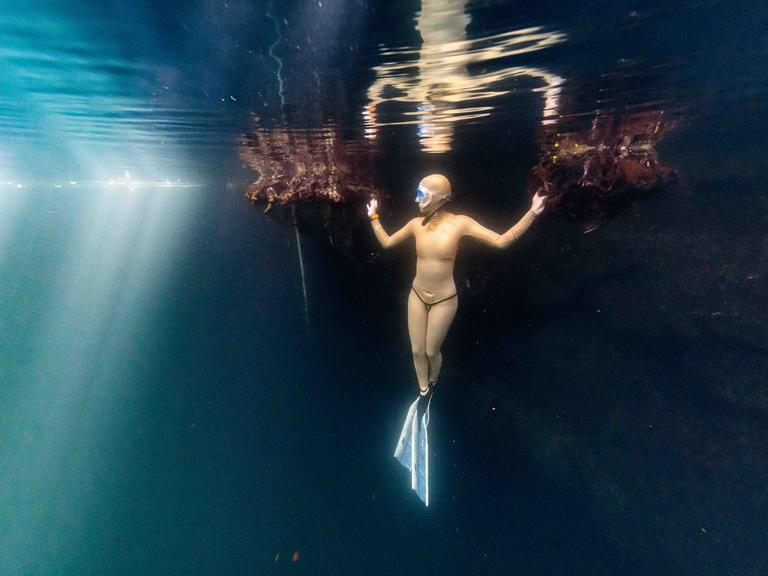 Eine Taucherin steht förmlich unter Wasser. Sie trägt einen hellorangen Tauchanzug und hält sich mit beiden Händen an undeutlichen, schwimmenden Strukturen fest.