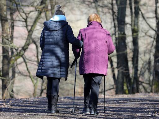 Eine alte Frau mit Stöcken wird von einer jüngeren beim Spazierengehen gestützt.