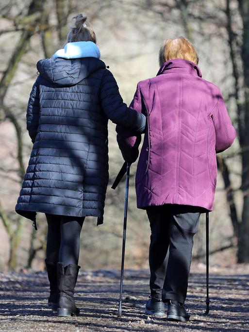 Eine alte Frau mit Stöcken wird von einer jüngeren beim Spazierengehen gestützt.