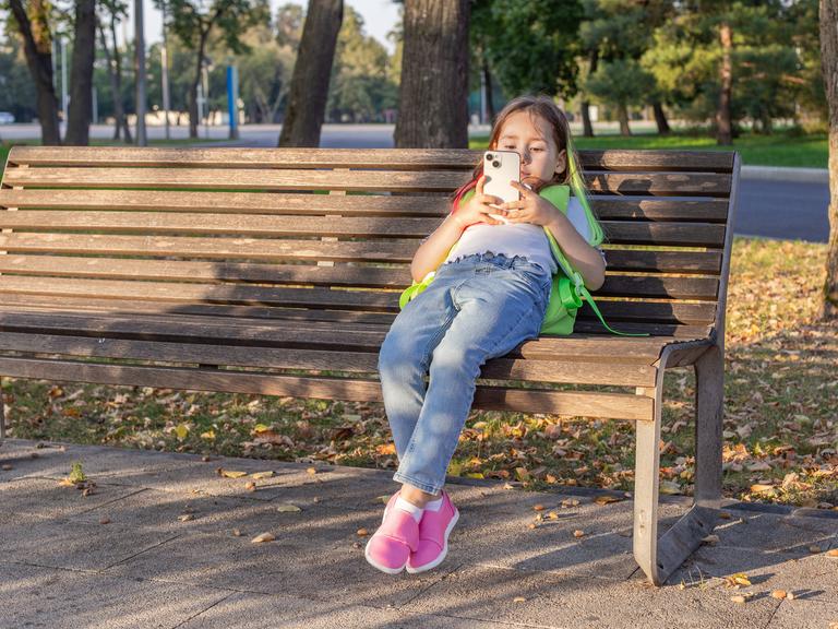 Junges Mädchen sitzt tief zurückgelehnt auf einer Parkbank und benutzt ihr Smartphone.