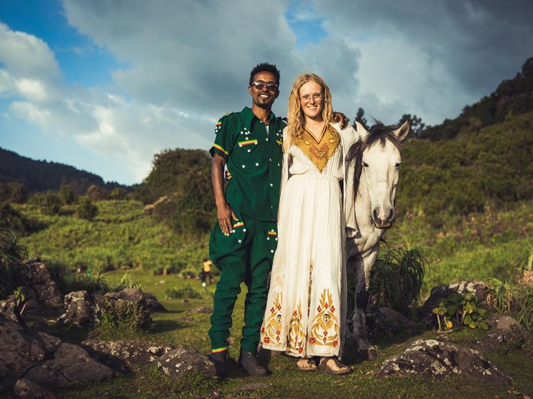 Valerie Seitz und ihre Mann Abiy stehen Arm in Arm zusammen mit ihrem Pferd in einem grünen Tal.